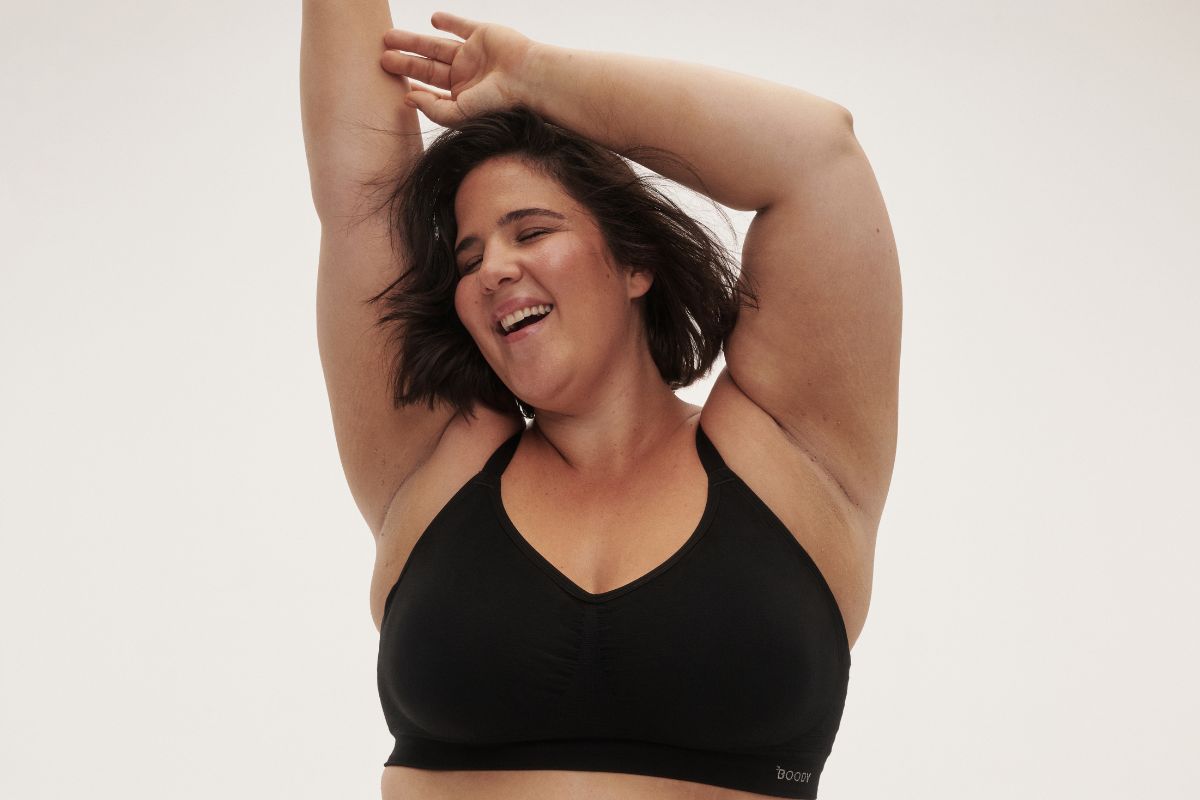 Smiling model wearing a black wireless bra with arms raised against a neutral studio backdrop.